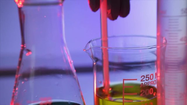 The camera tilts down from the gloved hand of a chemist or technician stirring liquid in a laboratory beaker.