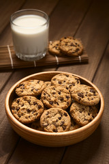 Chocolate Chip Cookies oder Kekse mit Schoko-Tröpfchen in Holzschüssel mit einem Glas Milch im Hintergrund (Selektiver Fokus, Fokus auf das mittlere Plätzchen)