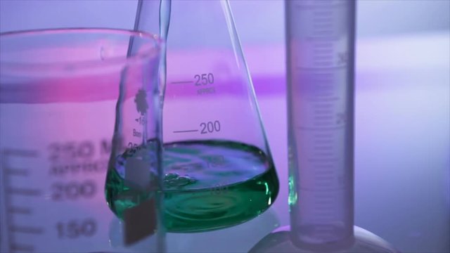 A green liquid pours into a flask in the background with an empty chemistry beaker and graduated cylinder out of focus in the foreground.