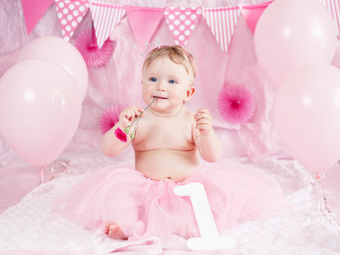 Portrait of cute adorable Caucasian baby girl with blue eyes in pink tutu skirt celebrating her first birthday with flower, gourmet cake and balloons looking away, cake smash first year concept