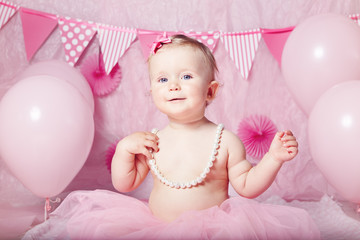 Portrait of cute adorable Caucasian baby girl with blue eyes in pink tutu skirt and pearls celebrating her first birthday with balloons looking in camera, cake smash first year concept