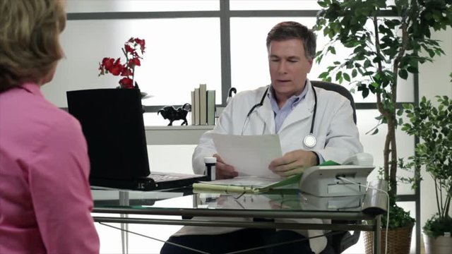 A doctor sitting in his office talks to a patient to whom he hands a prescription.