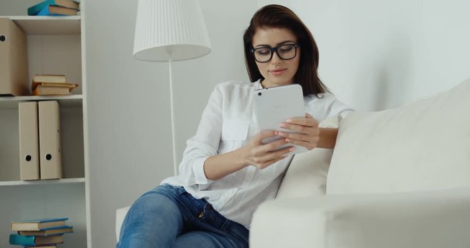 Young brunette businesswoman in white shirt, blue jeans and glasses with computer tablet siting on the white sofa.