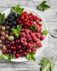 Assortment of berries - raspberries, gooseberries, red currants, cherries, black currants on a white plate on a light rustic wooden background. Healthy food, delicious summer dessert