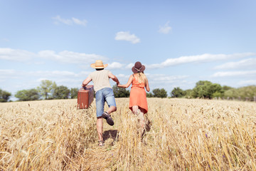 Naklejka na ściany i meble Couple running in field, man holding in his hand vintage suitcase on countryside landscape blue sky outdoors background