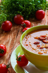 Soup: tomato soup with smoked sausage, tomatoes and lentil. Green bowl on wooden table. Small cherry tomatoes and green lettuce on table.