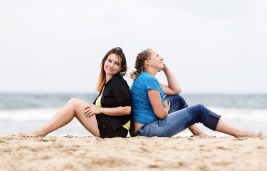 Fototapeta na wymiar Two pretty young beautiful smiling women sitting with his back to each other and resting on the sand on the seashore. Two girls resting on the beach. Two girls outdoors. Selective focus on the models.