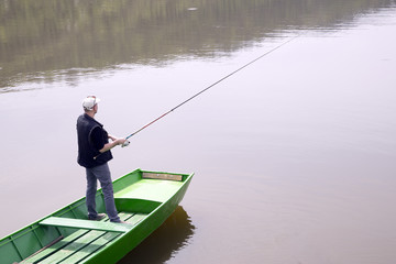 Fisherman Casting A Fishing Rod From The Green Boat On The Lake And Patiently Waiting For Fish To Take A Bait, Water Reflection