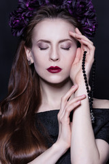 Attractive brunette woman with perfect dramatic makeup in a wreath of purple flowers. Her eyes closed. Her hair is shining, her skin glows. She holds a black rosary.