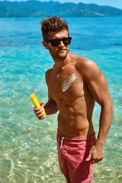 Sun Skin Protection In Summer. Handsome Man In Sunglasses Sunbathing With Sunscreen Lotion On Sexy Athletic Muscular Body. Male Fitness Model Tanning Using Solar Block Cream For Healthy Tan. Skincare 
