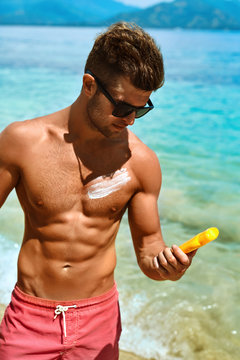 Summer Skin Care. Handsome Sexy Man In Fashionable Sunglasses Tanning Using Protective Sunscreen Lotion On Athletic Muscular Body. Male Model Sunbathing With Solar Sun Block Protection Cream On Beach