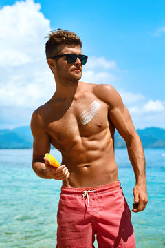 Summer Skin Care. Handsome Sexy Man In Fashionable Sunglasses Tanning Using Protective Sunscreen Lotion On Athletic Muscular Body. Male Model Sunbathing With Solar Sun Block Protection Cream On Beach