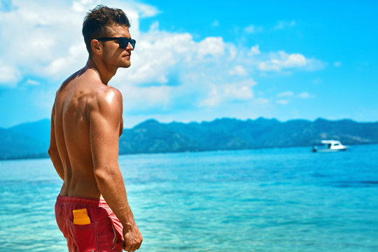 Summer Travel Vacation. Handsome Man With Sexy Body In Fashionable Sunglasses Sunbathing, Tanning At Sea Beach. Fitness Male Model With Sunscreen Lotion, Sun Block Skin Protection Cream In Pocket