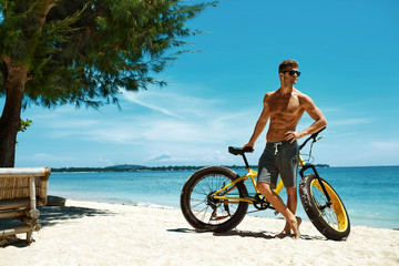 Fototapeta premium Handsome Muscular Man With Yellow Sand Bicycle Relaxing On Shore On Summer Travel Beach Vacation. Fitness Male Model With Bike Sunbathing By Ocean. Sports Activity And Recreation In Summertime Concept