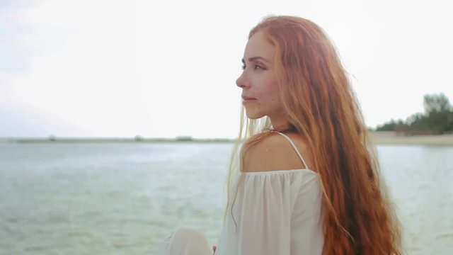 Attractive woman with long red hair at the beach looking to the camera and smiling. slow motion