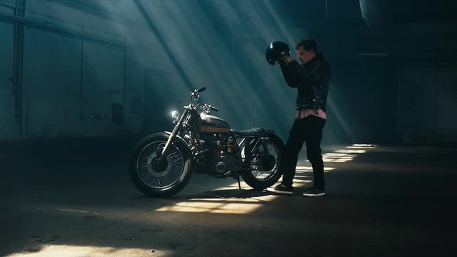 Young thoughtful Caucasian male biker in leather jacket mounts his custom cafe racer motorcycle in large warehouse garage. 60 FPS slow motion Blackmagic URSA Mini RAW graded footage