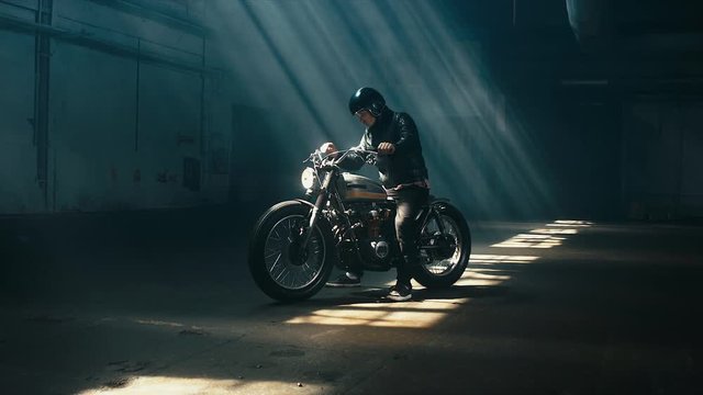 Young Caucasian male biker in leather jacket driving his custom cafe racer motorcycle in large warehouse garage. 60 FPS slow motion Blackmagic URSA Mini RAW graded footage