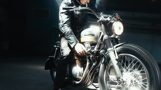 Close up shot of young Caucasian male biker in leather jacket riding his custom cafe racer motorcycle in large warehouse garage. 60 FPS slow motion Blackmagic URSA Mini RAW graded footage