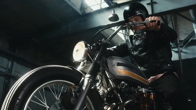 Close up shot of young Caucasian male biker in leather jacket riding his custom cafe racer motorcycle in large warehouse garage. 60 FPS slow motion Blackmagic URSA Mini RAW graded footage