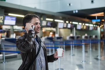 Portrait of young handsome man walking in modern airport terminal, talking smart phone, travelling with bag and coffee, wearing casual style clothes, blurred registration desks on background