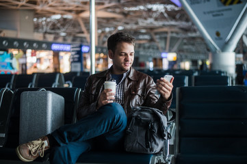 Handsome smiling man in casual wear holding luggage and messaging through his mobile phone while...
