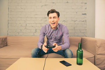 Excited  man sitting on sofa with bottle of beer and playing vid © deagreez