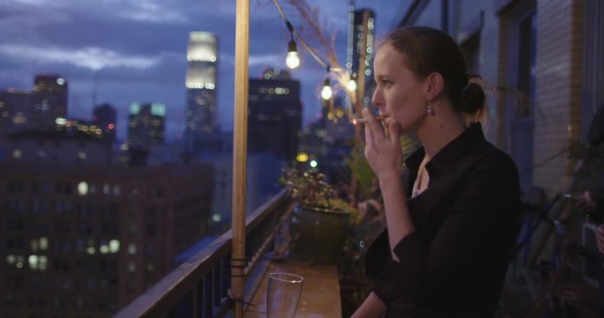 Attractive young woman smokes a cigarette in front of the night skyline from a balcony in Downtown Los Angeles, California.  Recorded in slow motion at 60fps.