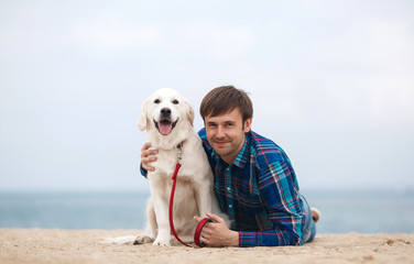 A nice young man,with a fashionable hairstyle,wearing a blue plaid shirt,spends time on the beach in the spring with his friend,a dog breed golden retriever lying on the sand against the blue ocean