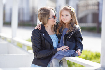 Fototapeta premium Beautiful woman in red mirrored sunglasses in the shape of hearts,a black leather jacket,spending time outdoors in the spring with his blond daughter,a girl of four years old,wearing a black jacket