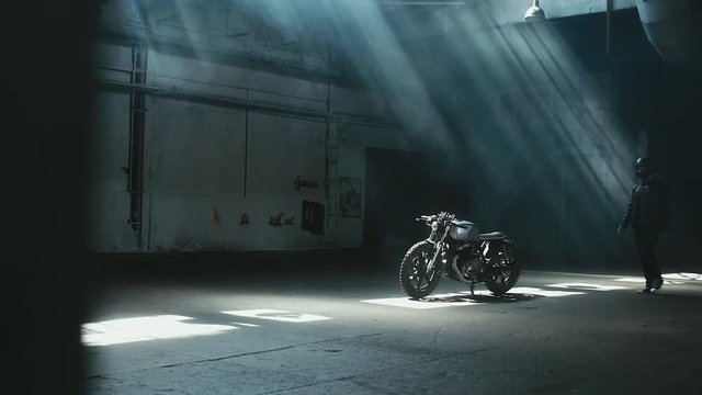 Young thoughtful Caucasian male biker in leather jacket mounts his custom cafe racer motorcycle in large warehouse garage. 60 FPS slow motion Blackmagic URSA Mini RAW graded footage
