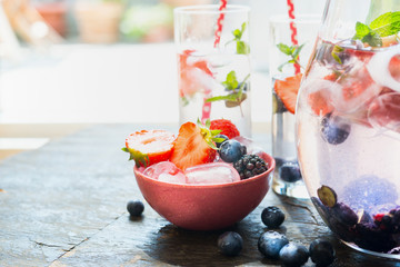 Ice cubes and berries in red bowl on table with glass of water. Summer drink preparation