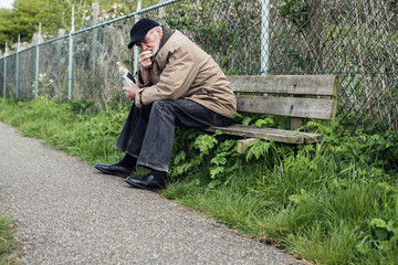 Senior homeless man on bench holding bottle.