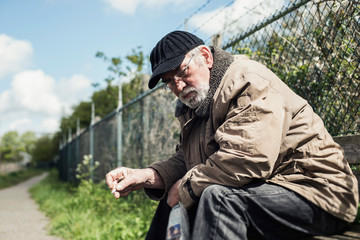 Homeless man on bench in park smoking cigarette.