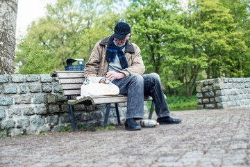 Homeless man searches bag on bench in park.