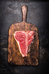 Raw T-bone steak on aged wooden cutting board on dark rust metal background, top view, close up