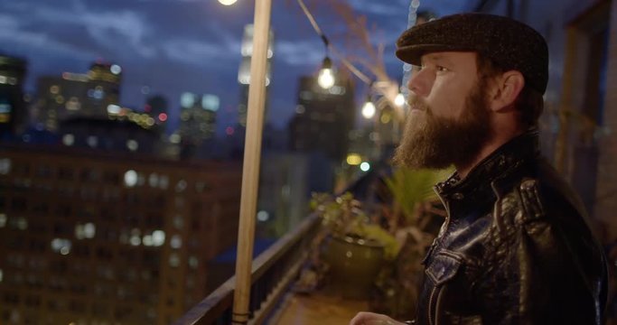 Bearded man in leather jacket and flat cap drinking beer alone on balcony at night in Downtown Los Angeles, California.  Recorded in slow motion at 60fps.