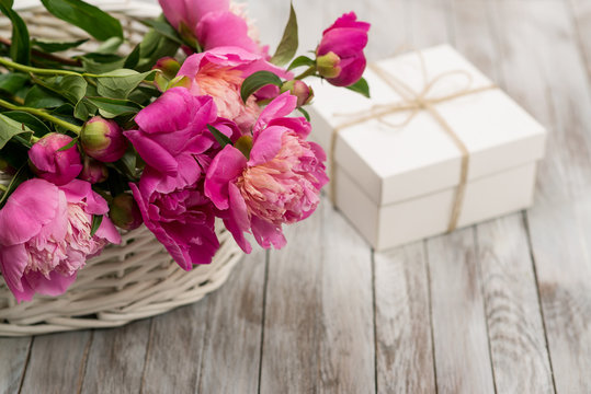 Beautiful flowers peonies in basket with gift box on light wooden background.