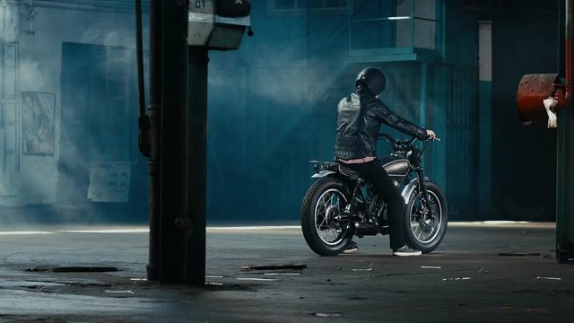 4K cinemagraph - young Caucasian male biker in leather jacket sitting on his custom cafe racer motorcycle in large warehouse garage looking forward. Background fog seamless loop