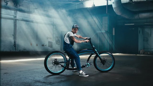 4K cinemagraph - young Caucasian male in denim overalls with tattoos sitting on his custom made bicycle in large warehouse garage looking into camera. Background fog seamless loop
