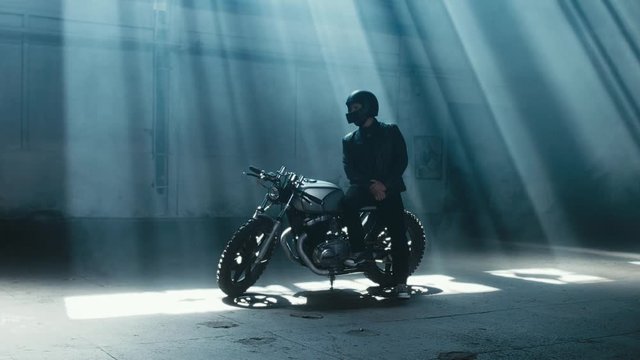 4K cinemagraph - young Caucasian male biker in leather jacket sitting on his custom cafe racer motorcycle in large warehouse garage looking forward. Background fog seamless loop