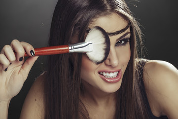 Young attractive playful young woman having fun while holding makeup brush over her eye over dark...