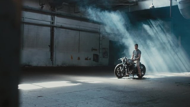 4K cinemagraph - young Caucasian male biker in shorts sitting on his custom cafe racer motorcycle in large warehouse garage looking forward. Background fog seamless loop