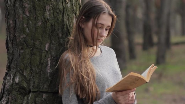 Teenage girl reading classic book leaning against tree trunk in forest in early spring, leisure activity studying outdoor nature park concept