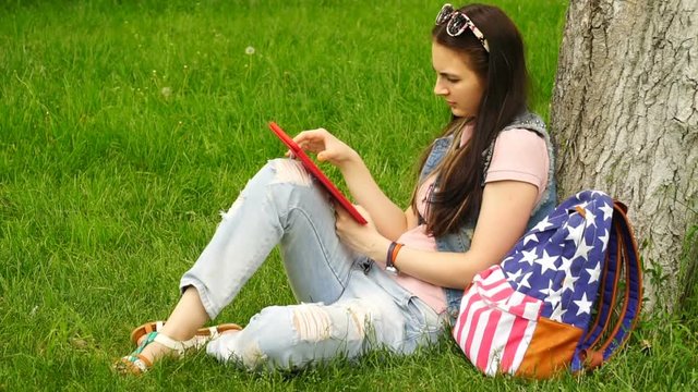 footage young beautiful woman sitting near a tree on the grass in the park and use the app in your comuter tablet.