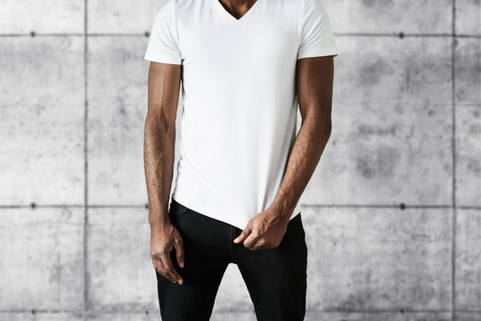 Cropped portrait of attractive African American man in trendy black jeans and white copy space T-shirt for your advertisement. Fit young black male posing against gray brick wall background