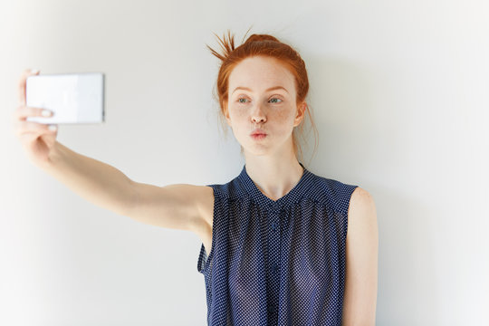 Portrait of beautiful redhead girl wearing spotted dress making self-portrait using cell phone, sending a kiss. Young female with freckles taking pictures against white wall. Selective focus