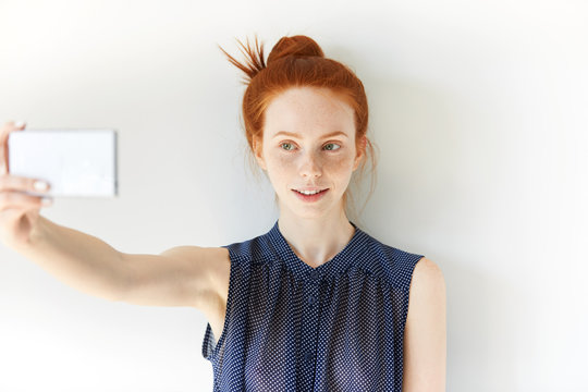 Smiling young woman making selfie using smart phone. Student girl with red hair and green eyes taking photograph with mobile phone, looking with pretty smile at the camera of electronic device