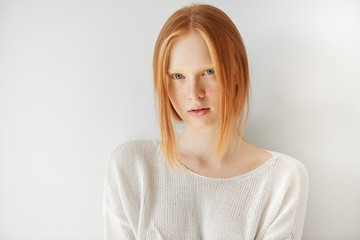 Beautiful female teenage model wearing casual white top looking at the camera with thoughtful expression on her face. Isolated portrait of shy redhead student girl with freckles and no make up