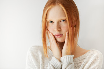 Close up shot of cute redhead teenage girl with perfect freckled skin and blue eyes wearing casual clothes, with hands on her cheeks, looking at the camera with serious expression on her face