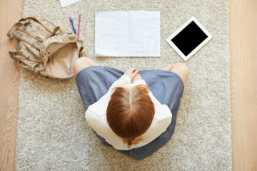 Overhead portrait of young female entrepreneur working on her business concept at home, sitting with crossed legs on the floor. Student girl with red hair preparing for exams using electronic gadget